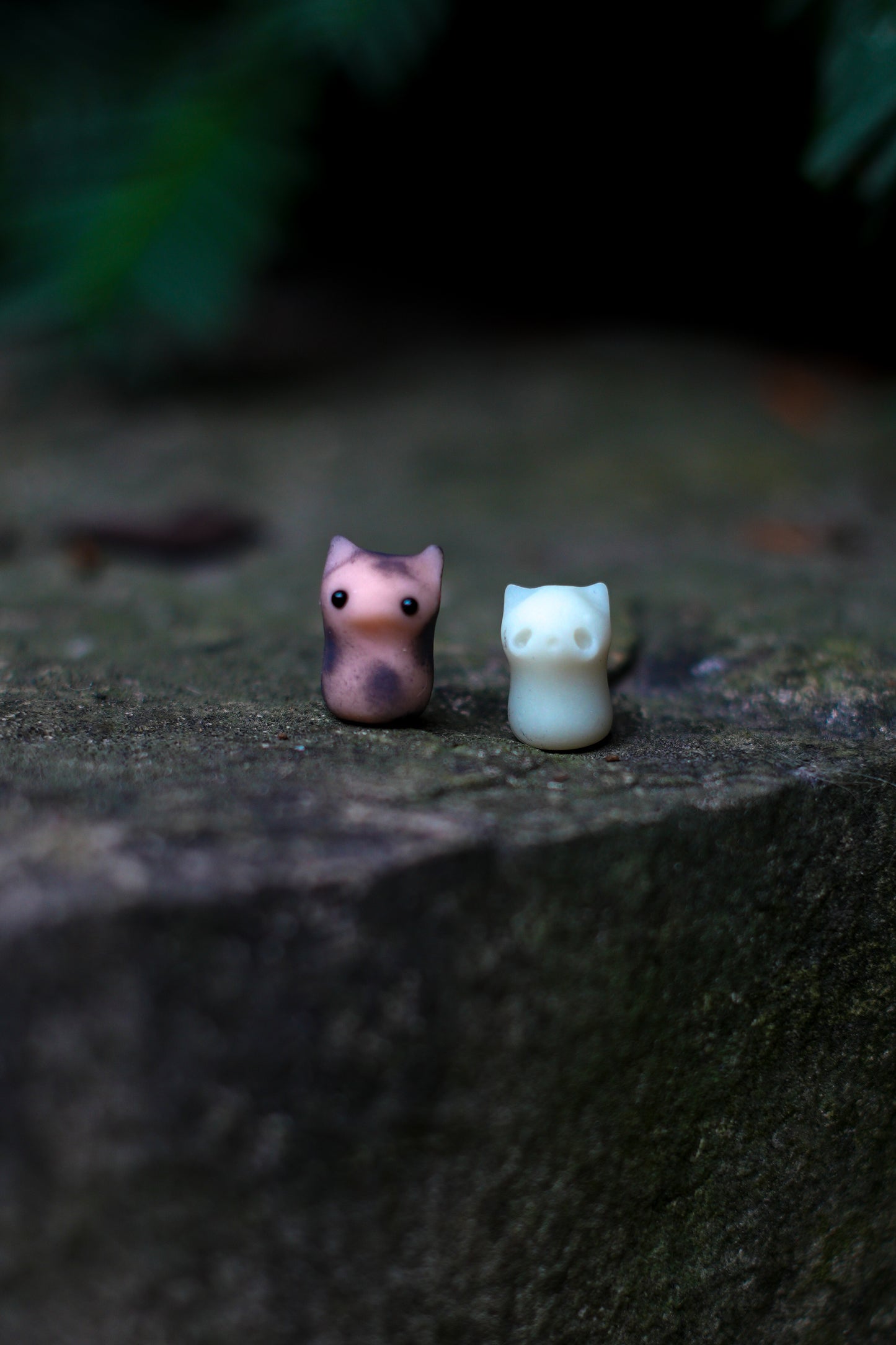 Two small figurines of a spotted kitten and a white ghost cat on a dark stone surface with blurred greenery in the background.
