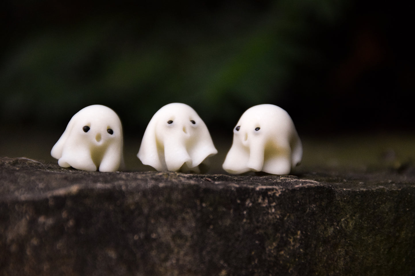 Three small white ghost figurines on a dark surface with a blurred natural background