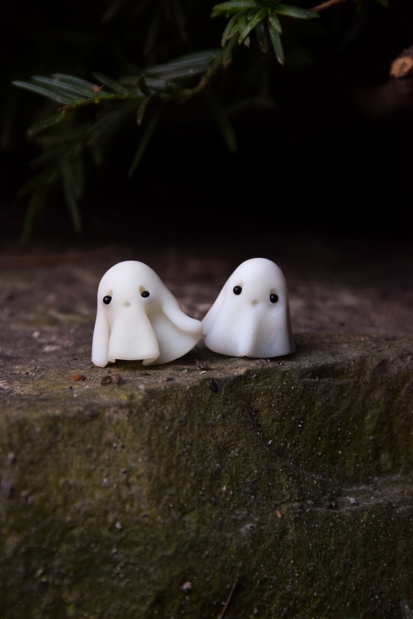 Two small white ghost figurines sitting on a stone surface with a dark background.