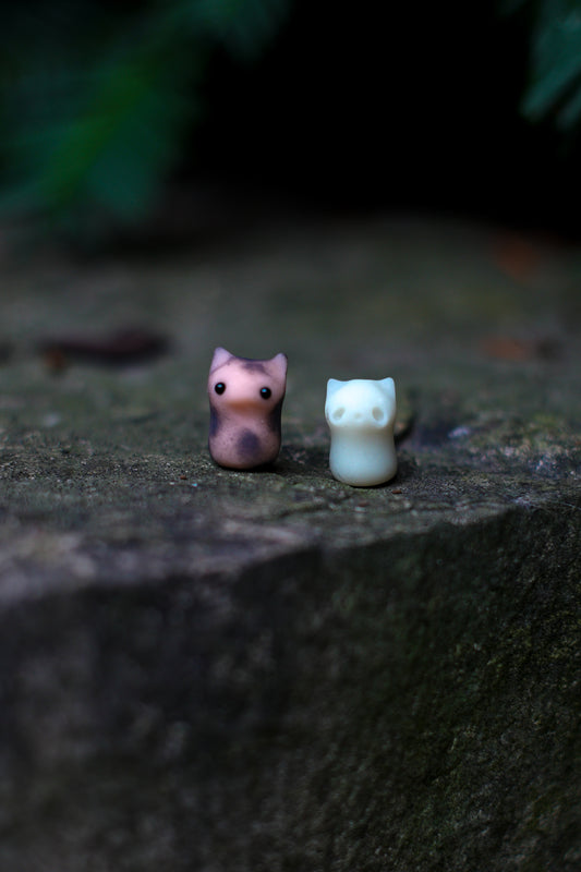 Two small figurines of a spotted kitten and a white ghost cat on a dark stone surface with blurred greenery in the background.